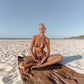 Woman in a bikini sitting on a towel on a sandy beach with a clear sky.