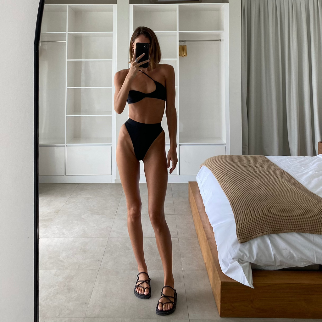 A person standing in front of a mirror wearing a black bikini top and matching bottoms, with the mirror reflecting a bedroom setting.