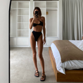 A person standing in front of a mirror wearing a black bikini top and matching bottoms, with the mirror reflecting a bedroom setting.