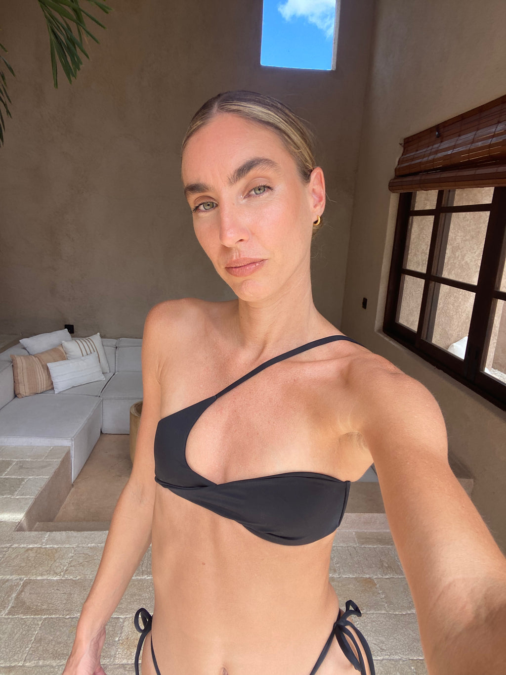 Woman wearing a black bikini standing in an outdoor setting with a neutral wall and window in the background.